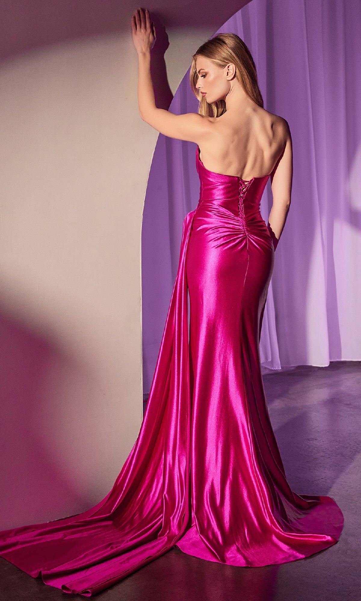 A woman in the Ladivine CD379 Side-Sash Strapless Long Prom Gown, featuring a shiny, bright pink fabric and long train, stands with her back to the camera, touching a wall in a softly lit room.